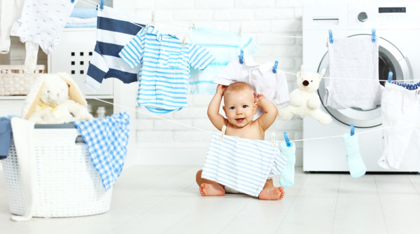 Một người mẹ đang ôm em bé sơ sinh mặc quần áo sạch sẽ, da dẻ hồng hào không nổi mẩn đỏ, bên cạnh là chai nước giặt hữu cơ cho bé chiết xuất thiên nhiên an toàn.